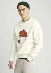 Sweatshirt de couleur crème avec un graphique d'une tasse remplie de fruits bruns et une fraise sur le côté inférieur. Poignets et col tricotés.