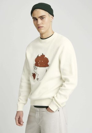Sweatshirt de couleur crème avec un graphique d'une tasse remplie de fruits bruns et une fraise sur le côté inférieur. Poignets et col tricotés.