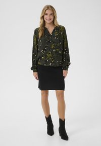 Junge Frau in grüner, blumiger Knopfleiste-Bluse, schwarzem knielangen Rock und schwarzen Stiefeletten, die vor einem schlichten Hintergrund steht.