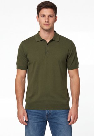 Giovane uomo che indossa una polo a maniche corte verde oliva e jeans blu, in piedi contro uno sfondo bianco uniforme.