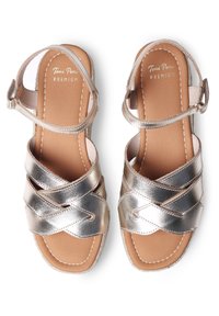 Sandalias metálicas plateadas con correas entrecruzadas, hebilla ajustable en el tobillo y plantilla beige. Textura suave y diseño de puntera cuadrada.