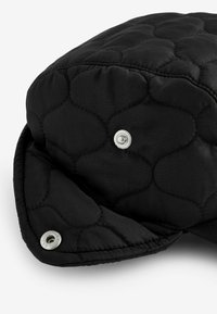 Chapeau noir matelassé avec une texture moelleuse. Présente des éléments en métal argenté et des bords arrondis, mettant en avant un motif en relief subtil.