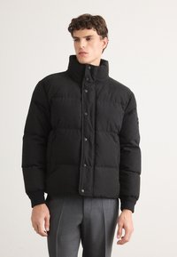 Geacă puf black cu guler înalt, butoni cu clapetă și buzunare pe laterale. Are o textură matlasată și manșete ribbate, purtată peste pantaloni gri.