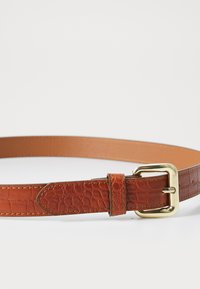 Bruine leren riem met krokodillenstructuur, voorzien van een gouden metalen gesp en een gladde binnenlaag. Inclusief een bijpassende leren houder.