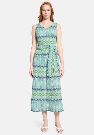 Frau trägt ärmellosen V-Ausschnitt-Overall mit grünem, blauem und schwarzem Zickzackmuster und passendem Stoffgürtel, steht vor weißem Hintergrund.