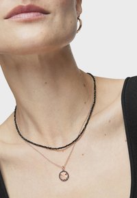 Dos collares en capas: uno con perlas negras, el otro en oro rosa con un colgante circular que tiene un contorno de gema. Texturas suaves.