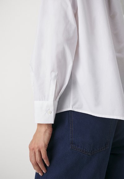 Weißes Hemd mit Knopfleiste und langen Ärmeln, die mit einer Manschette und Knopfdetails versehen sind, kombiniert mit dunkelblauen Jeans. Glatte Stoffoberfläche.
