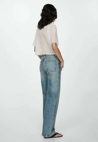 Haut blanc court avec des détails en dentelle sur les manches, associé à un jean large bleu délavé et des tongs, vu de dos.