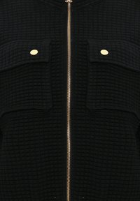 Tissu noir texturé avec fermeture éclair centrale dorée et deux poches à rabat sécurisées par de petits boutons ronds dorés.