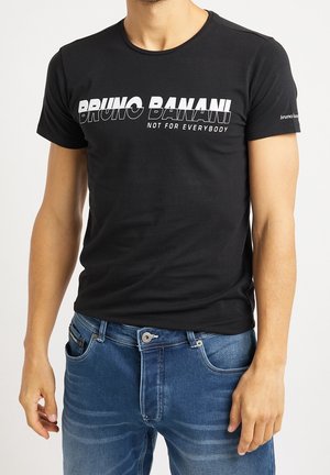 Mann trägt ein schwarzes Bruno Banani T-Shirt mit weißem Text "Nicht für jedermann" kombiniert mit blauer Jeans vor weißem Hintergrund.