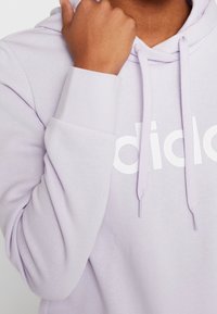 Felpa con cappuccio lavanda con logo bianco del brand, lacci e polsini a coste. Texture del tessuto morbida e design casual. Mano appoggiata vicino al mento.