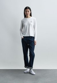 Γυναίκα που στέκεται απέναντι σε έναν απλό τοίχο, φορώντας λευκό πουκάμισο με μακριά μανίκια, σκούρα τζιν που έχουν γυριστεί και λευκά αθλητικά παπούτσια, με το ένα χέρι στην τσέπη.