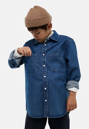 Chemise en denim bleu profond avec des manches longues, deux poches poitrine et des boutons-pression blancs. Associée à une chemise à manches longues de couleur claire en dessous.