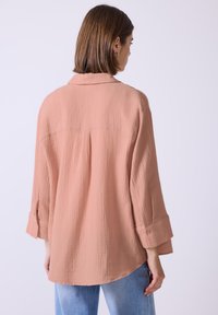 Camicia rosa chiaro, morbida e tessuta, con colletto, maniche arrotolabili e dettagli con bottoni. Pinces sul retro per un tocco di design in più.
