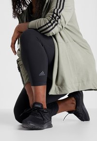 Veste verte zippée avec des rayures noires, leggings noirs et chaussures de sport noires à semelles texturées, avec le logo Adidas.