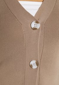 Närbild av en beige ribbad stickad cardigan med två stora vita och bruna marmorliknande knappar knäppta över en vit undertröja.