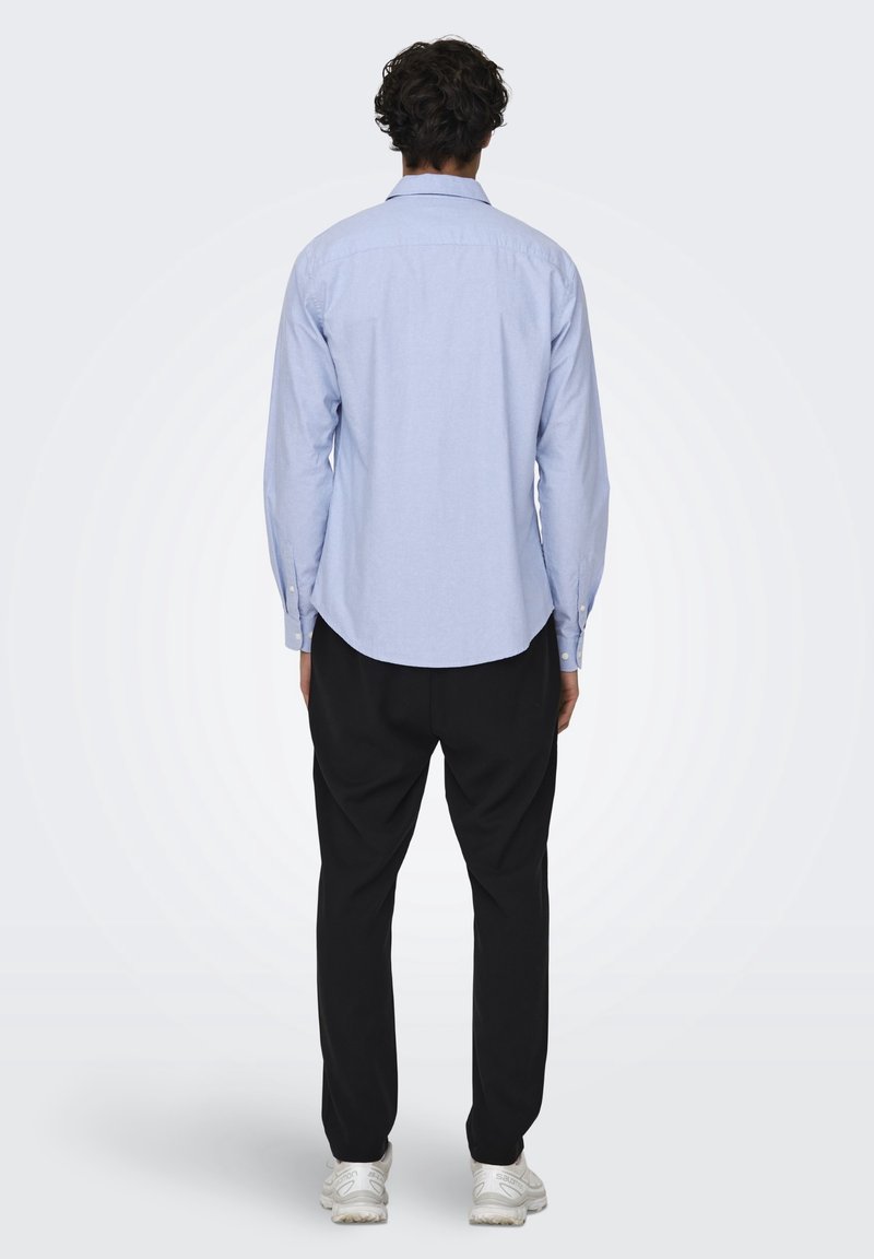 Hombre de pie de espaldas, vestido con una camisa de manga larga azul clara, pantalones negros y zapatillas blancas, sobre un fondo liso.