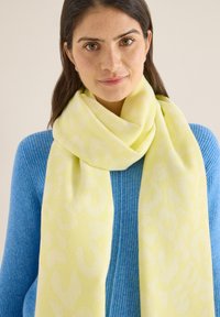 Écharpe jaune clair à motif léopard subtil, fabriquée en matériau doux, drapée autour d'un pull bleu. Texture lisse avec des bords arrondis.