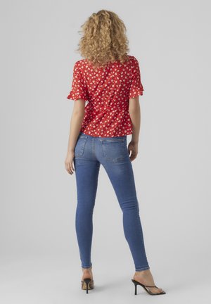 Žena s kudrnatými blond vlasy, oblečená v červené květované blůze, modrých úzkých džínách a černých sandálech na podpatku, stojící zády.