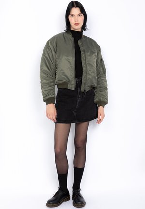 Giacca bomber verde con finitura lucida, dolcevita nera, gonna di jeans nera, collant neri trasparenti e scarpe stringate nere.