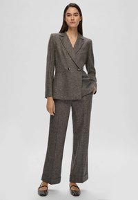 Donna in piedi che indossa un blazer grigio doppiopetto e pantaloni a gamba larga coordinati, con scarpe flat a motivi bianco e nero.