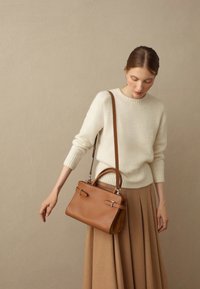 Borsa a tracolla in pelle color tan con doppi manici e tracolla rimovibile. Il modello indossa un maglione color crema e una gonna plissettata beige su uno sfondo neutro.