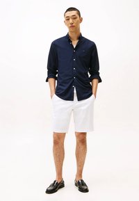 Camicia blu con bottoni, pantaloni corti bianchi e mocassini neri. La camicia ha maniche lunghe e i pantaloni corti sono fino al ginocchio. Tessuto con una texture liscia.