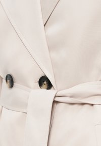 Beige περιτύλιγμα μπουφάν με υφή, διακοσμημένο με μαύρους κουμπιά και ζώνη που δένει μόνο του. Ο σχεδιασμός περιλαμβάνει βαθύ V-ντεκολτέ και κομψά πέτα.