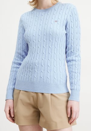 Femme portant un pull en grosse maille bleu clair et un short beige, debout devant un fond blanc uni.