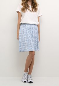 Vaaleansininen kuvioitu hame, jossa on kerätty vyötärö, yhdistettynä yksinkertaiseen valkoiseen t-paitaan ja harmaisiin lenkkareihin. Sileä kangas ja levenevä muotoilu.