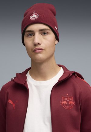 Młody mężczyzna w bordowej czapce FC Liefering i kurtce Red Bull Salzburg na białej koszuli, patrzący przed siebie z neutralnym wyrazem twarzy.
