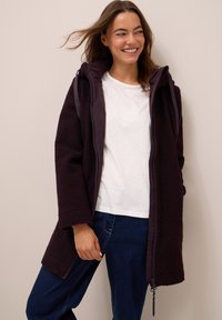Manteau en polaire violet avec une fermeture éclair à l'avant, grande capuche et poches latérales, porté sur une chemise blanche et un jean bleu foncé.