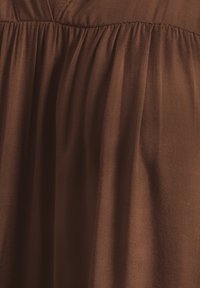 Brunt stof med en glat tekstur, der har sammenfoldede plisseringer øverst. Materialet har en let glans, hvilket skaber bløde draperinger.