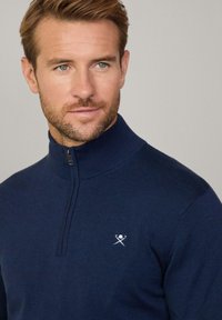 Sudadera de cremallera azul marino con cuello alto, textura acanalada y pequeño logo blanco con palos cruzados en el pecho.