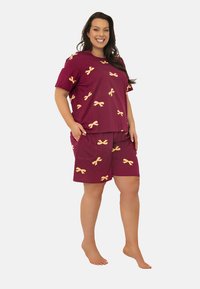 Uśmiechnięta kobieta stoi boso, ubrana w bordową koszulkę z krótkim rękawem i szorty w żółte wzory wstążek, z rękami w kieszeniach, na białym tle.