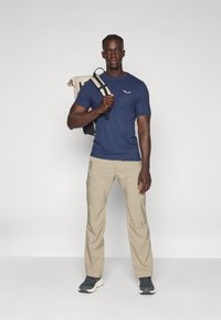 Tumesinise lühikese varrukaga särk logo ja khaki cargo püksid, samuti hallid tenniseketid. Mudel kannab beeži seljakotti mustade rihmadega.