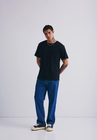 T-shirt nera in cotone con taschino, abbinata a jeans in denim blu a vestibilità ampia e sneakers nere con dettagli crema. Collana semplice a catena.