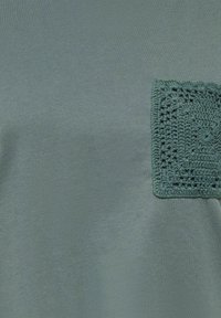 Grønt stof med en firkantet hæklet lap-pung med et detaljeret blomstermønster syet på øverste højre side.