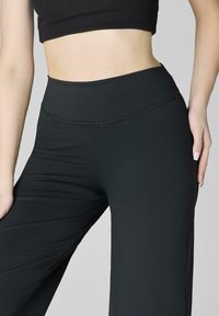 Kobieta w czarnych legginsach z wysokim stanem i czarnym cropped topem na jednolitym jasnoszarym tle.