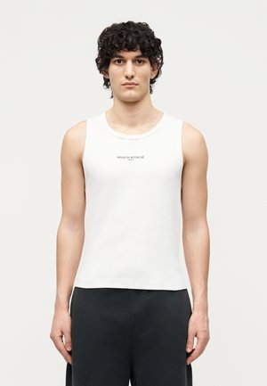 Jeune homme aux cheveux bouclés porte un débardeur blanc avec le logo "Maison Kitsuné Paris", associé à un pantalon noir, debout devant un fond uni.