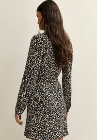 Vrouw met lang bruin haar, gekleed in een lange mouw zwarte en beige jurk met luipaardprint, staand met haar rug naar de camera gekeerd.