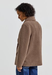 Kvinna med kort lockigt hår som bär en brun fleecejacka och blå jeans, ser ner och står mot en enkel ljus bakgrund.