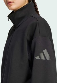 Black zip-up jacket with high collar, made from soft fabric. Features a reflective three-stripe design on the left sleeve.