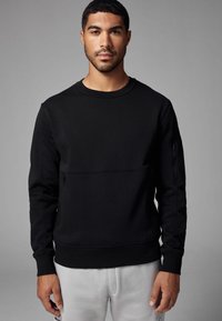 Sort sweatshirt lavet af blødt stof, med en rund hals, lange ærmer og en lynlåslomme på venstre arm.