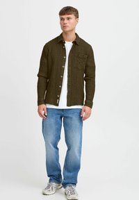 Camisa abotonada verde oliva con patrón texturizado, llevada sobre una camiseta blanca. Combinada con jeans azules y zapatillas de color claro.