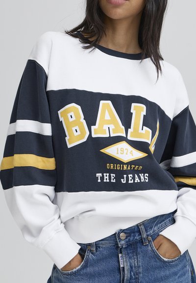 Sweatshirt alb și bleumarin cu accente aurii. Prezintă textul îndrăzneț "BALL", dungi orizontale pe mâneci și manșete ribbate. Purtați cu blugi albaștri.