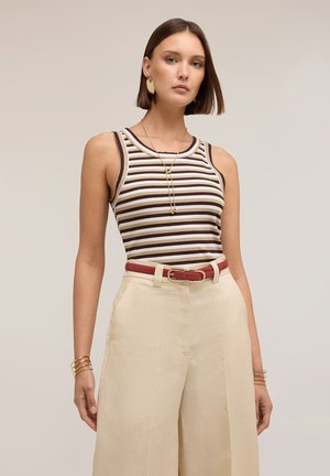 Femme portant un haut sans manches rayé marron et blanc, un pantalon beige taille haute avec une ceinture rouge, des boucles d'oreilles pendantes en or, un bracelet et un collier, posant.