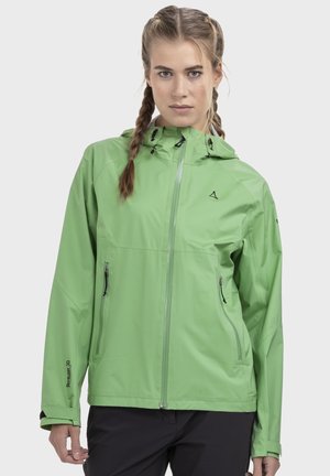 Frau mit geflochtenem Haar, die eine hellgrüne wasserdichte Kapuzenjacke und schwarze Hose vor einem einfarbigen Hintergrund trägt.