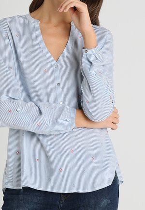 Femme portant un chemisier à rayures bleu clair et blanc à col en V avec de petits motifs cœurs roses et manches longues, associé à un jean bleu foncé.