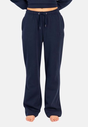 Mørkeblå, løse treningsbukser med elastisk midje og snor, brodert logo på venstre lår, brukt barbeint på lys bakgrunn.